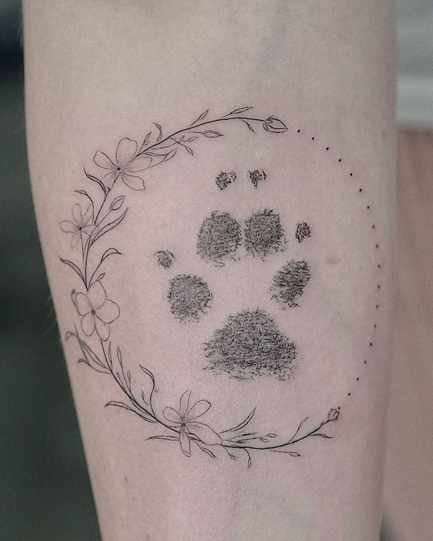 Empreinte de patte en pointillé dense, dans une couronne de fleurs en fine line refermée par un arc de points, sur l’avant-bras