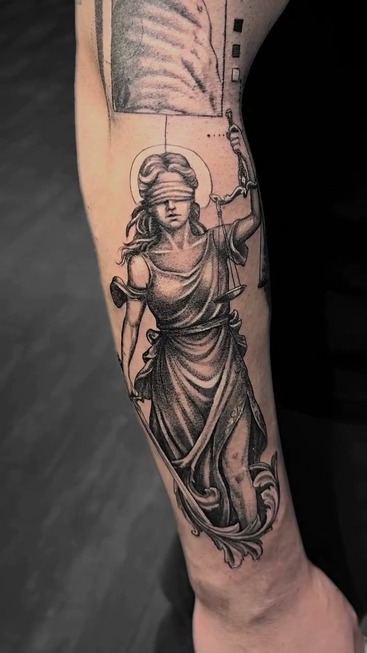 Statue de la Justice aux yeux bandés, auréolée, levant la balance et tenant l’épée, en réalisme noir et gris sur l’avant-bras ; au-dessus, le début d’une autre pièce de la manche