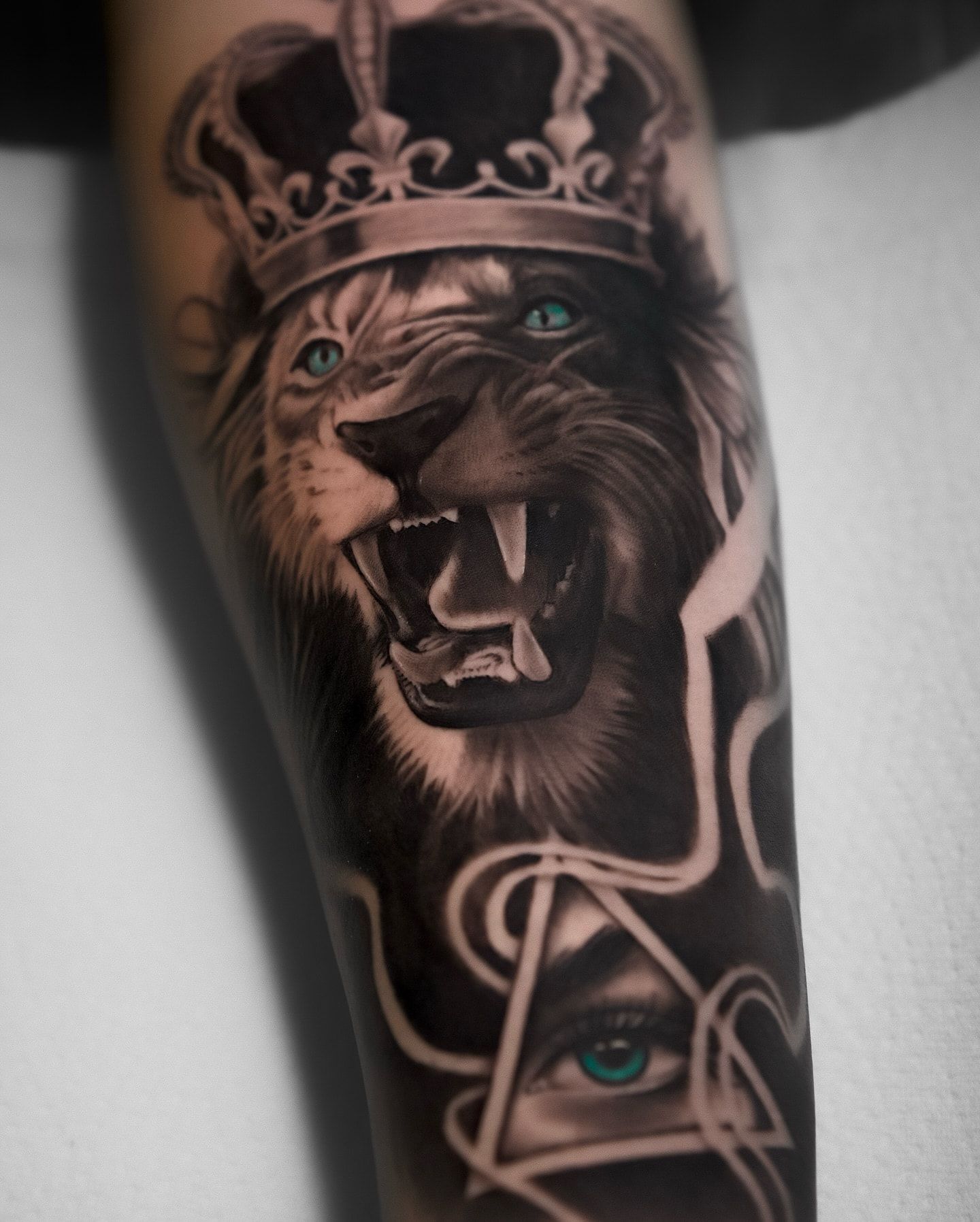León rugiente con corona y ojos turquesa sobre un ojo dentro de un triángulo, realismo en negro y gris en el antebrazo