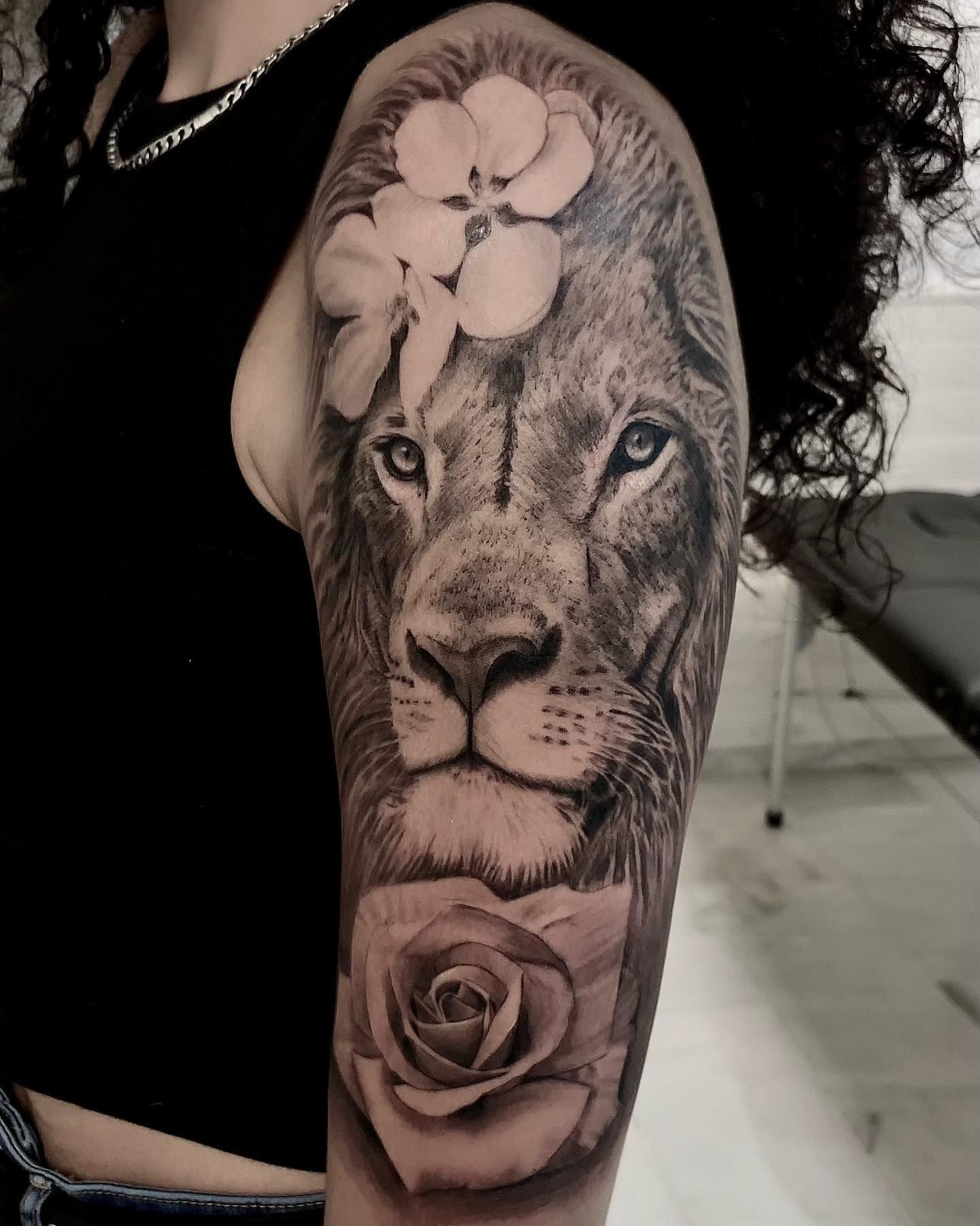 León de mirada serena adornado con orquídeas blancas sobre una rosa, realismo en negro y gris en el hombro y el brazo