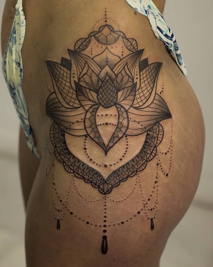 Fleur de lotus aux pétales en résille, bordure de dentelle et guirlandes de points terminées par des gouttes, tatouée en noir sur la hanche et le haut de la cuisse