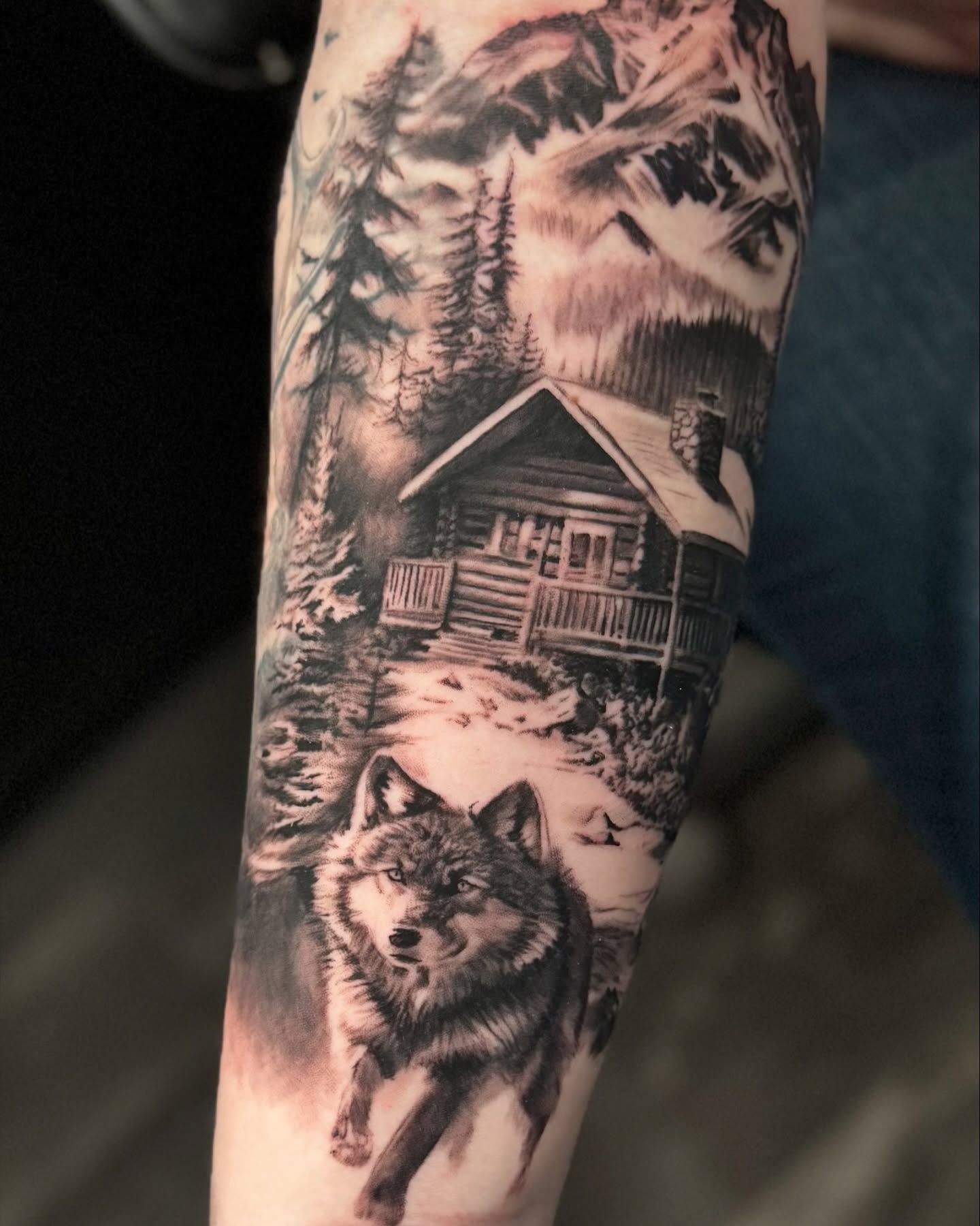Lobo que avanza ante una cabaña de troncos nevada, entre abetos y cumbres, realismo en negro y gris en el antebrazo