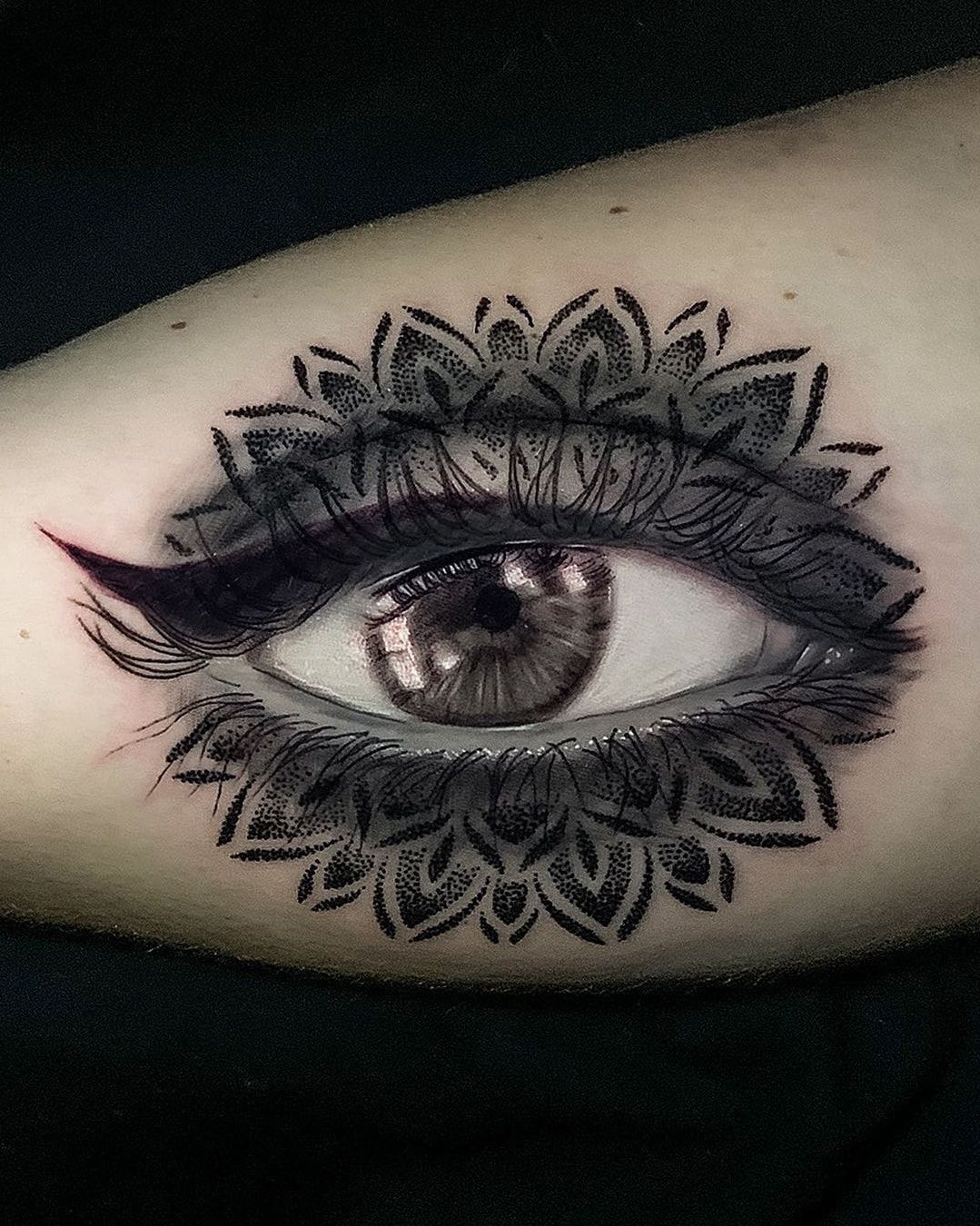 Œil réaliste souligné d’un trait d’eye-liner, encadré d’un mandala en pointillé, en noir et gris sur l’avant-bras
