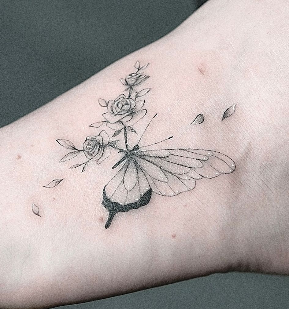 Papillon d’où naît une branche de roses, pétales envolés, en fine line aux ombrages délicats sur le dessus du pied