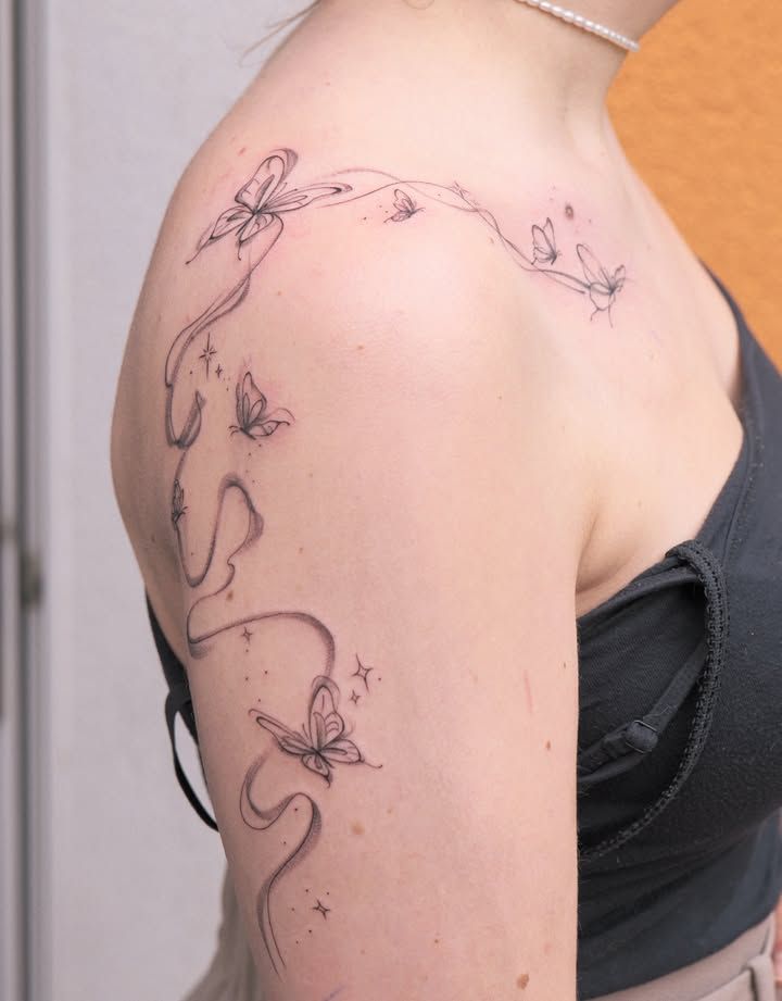 Papillons au trait fin reliés par un ruban ondulant et de petites étoiles, de la clavicule à l’épaule puis le long du haut du bras