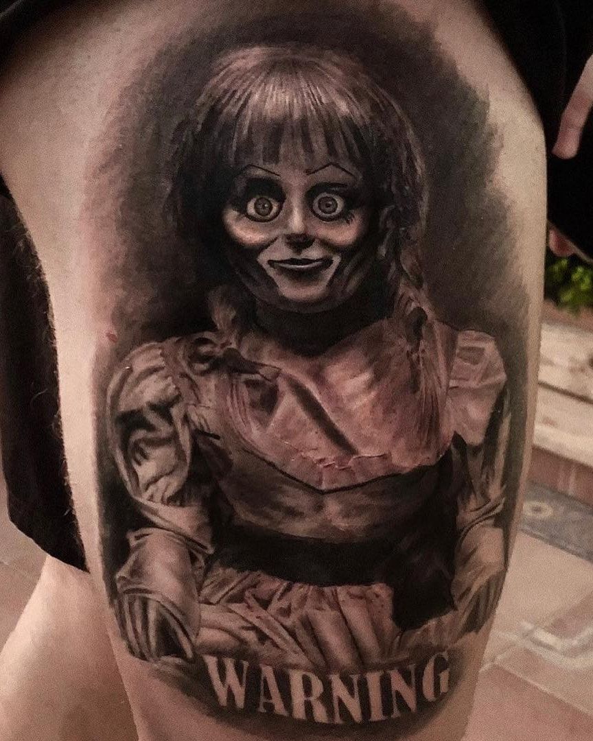 Poupée Annabelle au regard fixe et sourire figé au-dessus du mot WARNING, réalisme sombre en noir et gris sur la cuisse