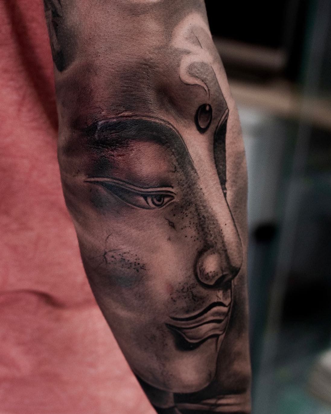 Visage de Bouddha serein aux yeux mi-clos, texture de pierre et volutes de fumée, réalisme noir et gris sur l’avant-bras